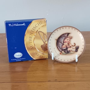 May include: A decorative plate depicting a child under an umbrella, alongside a blue box. The plate has a brown border with star details. The box is labelled "M.J. Hummel" and "Goebel Porzellanfabrik Germany".