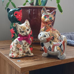 May include: Four decorative cat ornaments in different styles and colours. One cat has a floral pattern with a pink bow, another is green with red accents, and the others have unique patterns. They are displayed on a wooden surface.