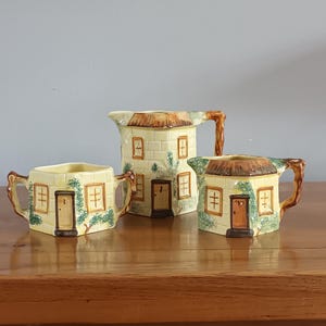 Puede incluir: Un juego de té de cerámica de tres piezas con un diseño de casa de campo. El juego incluye una crema, un azucarero y una tetera. Cada pieza está pintada de amarillo con detalles marrones, con ventanas, puertas y un tejado de paja.