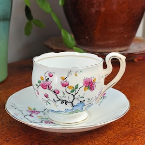 May include: A white teacup and saucer with a floral pattern. The cup has a scalloped rim and a handle. The saucer has a matching floral pattern. The flowers are pink, yellow, and green.