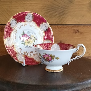 Puede incluir: Un juego de taza y platillo de té vintage. La taza blanca presenta un diseño floral con flores rosas y amarillas, y un borde dorado. El platillo y el interior de la taza son de color rojo intenso con un patrón blanco y dorado.