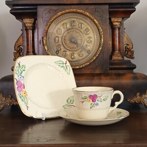 Puede incluir: Un juego de taza de té y platillo de cerámica color crema junto a un plato cuadrado a juego. Cada pieza presenta una delicada textura en relieve y un motivo floral pintado a mano en tonos morado, amarillo, azul y verde, con borde dorado.