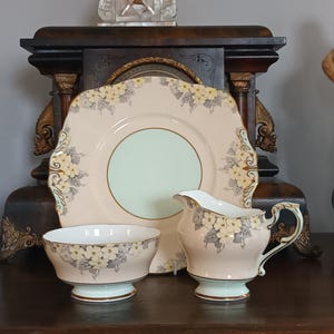 Può includere: Un servizio da tè vintage con un piatto quadrato, una lattiera e una piccola ciotola. Il set ha una base rosa pallido con un motivo floreale giallo e grigio. Il piatto e la ciotola hanno un centro verde menta.