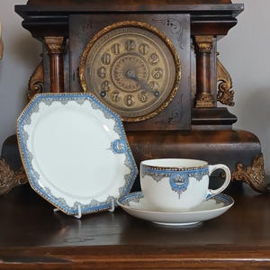 May include: A vintage tea set with a white and blue design. The set includes a teacup, saucer, and a decorative plate. The background features a dark wooden clock with a gold-coloured clock face.