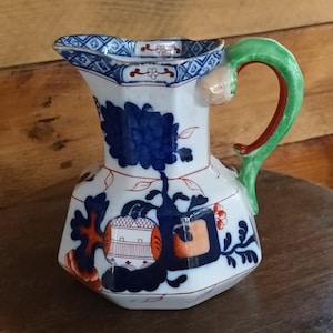 Puede incluir: Una jarra de cerámica blanca con un diseño floral azul y un asa verde. La jarra tiene forma hexagonal y está decorada con detalles azules, naranjas y rojos. El borde presenta un patrón geométrico azul y blanco.