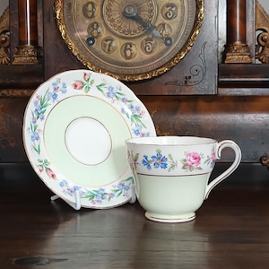 May include: A vintage teacup and saucer set with a light green base and floral detailing. The cup and saucer have a delicate design with blue and pink flowers. The set is placed on a dark wooden surface, with a clock in the background.