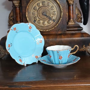 May include: A light blue porcelain tea set consisting of a teacup and two saucers, each featuring a scalloped edge, gold trim, and a delicate pattern of small orange flowers with green stems. The teacup has a gold-colored handle.