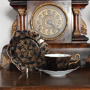 May include: A black and gold floral teacup and saucer set with a matching plate. The teacup has a gold handle and rim. The plate and saucer have a scalloped edge. A vintage clock is in the background.