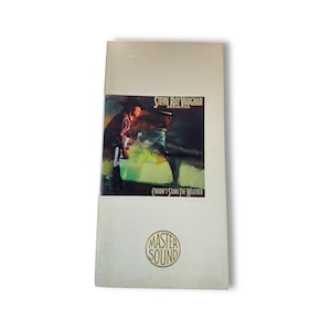 Puede incluir: Una funda de CD vintage con la carátula del álbum "Couldn't Stand the Weather" de Stevie Ray Vaughan. La portada presenta una foto de Vaughan tocando la guitarra. La funda es de color blanquecino con "Master Sound" impreso en la parte inferior.