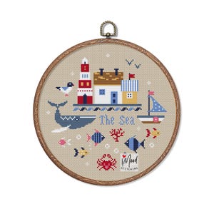 May include: A round embroidery hoop with a beige fabric background featuring a colorful cross-stitch design of a seaside scene. The design includes a blue whale, a lighthouse, a house, a sailboat, and the text "The Sea".