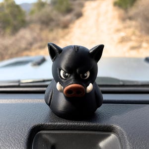 May include: A black figurine of a boar with an angry expression. The boar has white tusks, a brown snout, and white eyes. The figurine is sitting on a black dashboard.