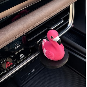 May include: A bright pink flamingo figurine with a white and black beak attached to a circular black base, mounted onto a car air vent. The flamingo has a rounded body and detailed wing textures, positioned against a dark vehicle dashboard interior.