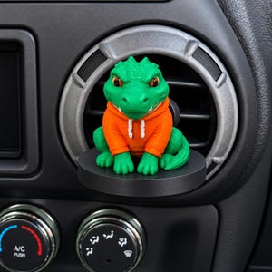 May include: A green lizard figurine wearing an orange hooded sweatshirt with white drawstrings, mounted on a circular black base attached to a circular car air conditioning vent. The lizard has textured skin and orange eyes, facing forward.
