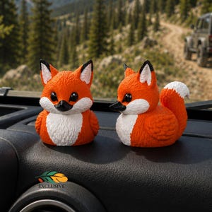 May include: Two orange and white fox-shaped rubber ducks with black eyes and noses. One duck faces forward while the other is in profile, showing a bushy tail. The figures are positioned on a black textured dashboard with a small logo below.
