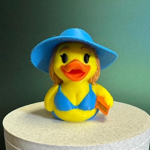 May include: A yellow rubber duck wearing a blue sun hat and bikini. The duck has an orange beak and is holding a small orange item. The duck is on a light gray surface with a dark green background.