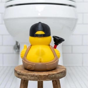 May include: A yellow rubber duck figure wearing a black baseball cap and brown overalls, holding a small black and red plunger. The duck is positioned on a small wooden stool. The figure features a small leaf emblem on the side of the overalls.
