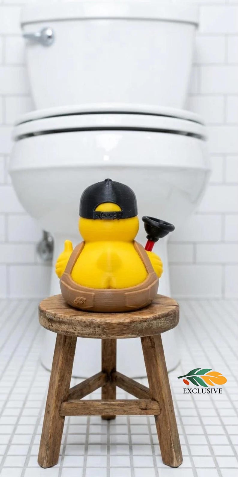 May include: A yellow rubber duck figure wearing a black baseball cap and brown overalls, holding a small black and red plunger. The duck is positioned on a small wooden stool. The figure features a small leaf emblem on the side of the overalls.
