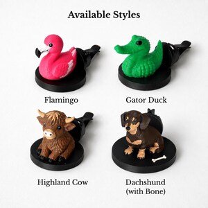 May include: Four car air freshener vent clips featuring animal figures on black circular bases. The styles include a bright pink flamingo, a green alligator-shaped duck, a brown Highland cow, and a brown and black dachshund dog with a small white bone.
