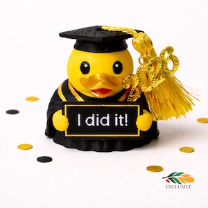 May include: A yellow graduation duck figurine wearing a black cap and gown, holding a sign that reads "I did it!". The duck has a gold tassel and the year "2023" attached. Gold and black confetti is scattered around the duck.