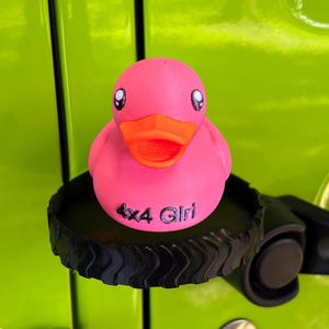 May include: A pink rubber duck with an orange beak and large eyes sits on a black tire-shaped base. The duck has the text "4x4 Girl" printed on its body. The background is a bright green surface.