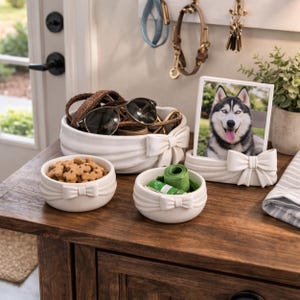 May include: A collection of white ceramic bowls and a picture frame, all adorned with bow accents. The bowls contain dog treats, sunglasses, and other items. A framed photo of a husky dog is also present. The items are arranged on a wooden surface.