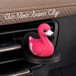 May include: A bright pink flamingo air vent scent clip featuring a white and black beak, mounted on a circular black base. The text Air Vent Scent Clip is displayed in white cursive script above the flamingo figure, which is attached to a car vent.