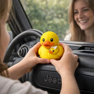 Puede incluir: Un pato de goma amarillo con un patito m&aacute;s peque&ntilde;o. Los patos tienen ojos negros y picos naranjas. El pato m&aacute;s grande tiene un lazo blanco. Los patos se sostienen frente al salpicadero de un coche.