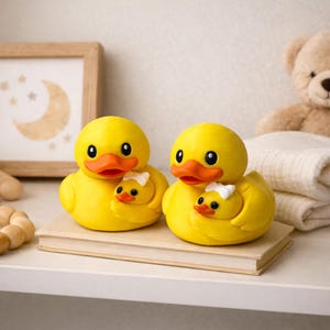 Puede incluir: Dos figuras de patos amarillos, cada una sosteniendo un patito m&aacute;s peque&ntilde;o. Los patos tienen picos naranjas y ojos negros. Los patitos tienen lazos blancos. Las figuras est&aacute;n sobre un libro, con un cuadro enmarcado y un oso de peluche al fondo.