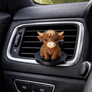 May include: A brown Highland cow figurine air freshener clip attached to a black car air vent. The cow has a textured coat, light tan horns, large dark eyes, and a white muzzle. It sits on a circular black base mounted to the horizontal vent slats.