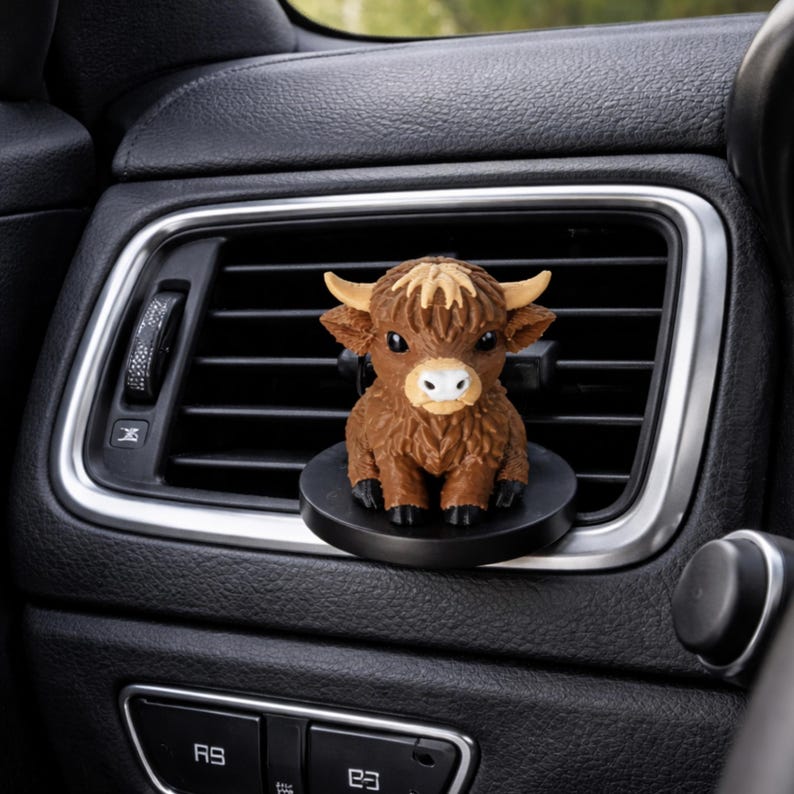 May include: A brown Highland cow figurine air freshener clip attached to a black car air vent. The cow has a textured coat, light tan horns, large dark eyes, and a white muzzle. It sits on a circular black base mounted to the horizontal vent slats.