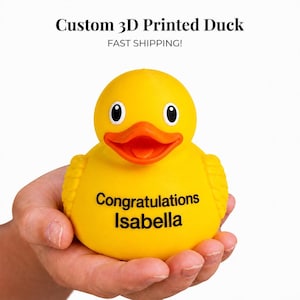 Peut inclure: Un canard en caoutchouc jaune vif imprimé en 3D avec un bec orange éclatant et des yeux noirs. L'avant du canard porte le texte noir Congratulations Isabella. Le canard présente un corps lisse et arrondi avec des détails d'ailes texturés.