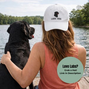 May include: White baseball cap with a black Labrador Retriever graphic and the text "LAKE DOG". A black Labrador sits next to a person wearing the hat. The image includes the text "Love Labs? Grab a Hat! Link in Description".