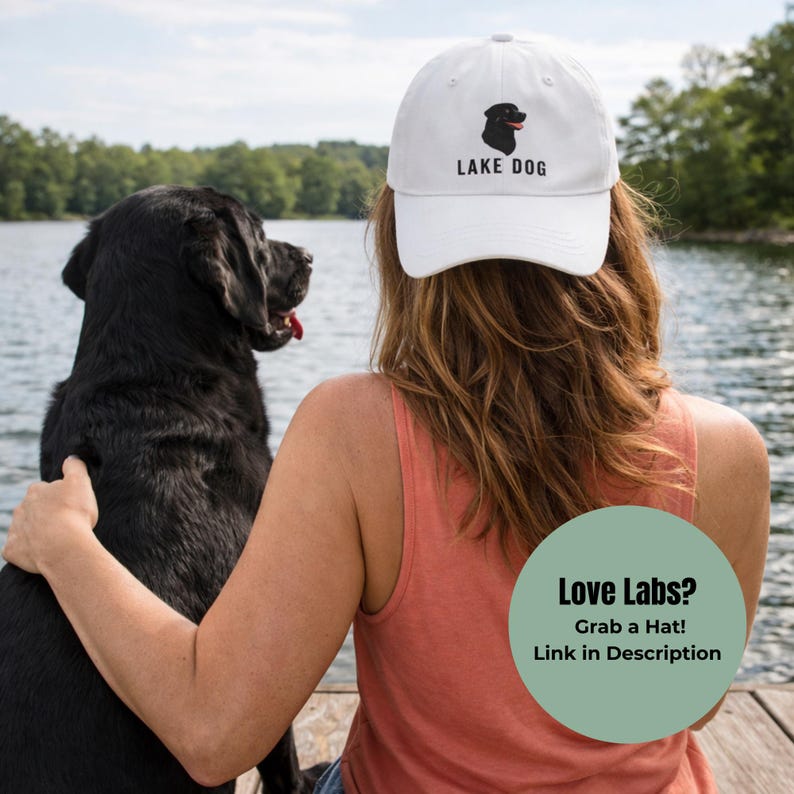 May include: White baseball cap with a black Labrador Retriever graphic and the text "LAKE DOG". A black Labrador sits next to a person wearing the hat. The image includes the text "Love Labs? Grab a Hat! Link in Description".