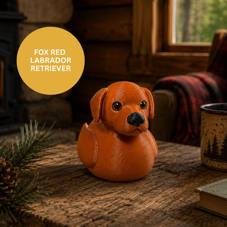 May include: A fox red Labrador Retriever figurine with a black nose and dark eyes. The figurine is sitting on a wooden surface. A yellow circle with the text "FOX RED LABRADOR RETRIEVER" is in the background.
