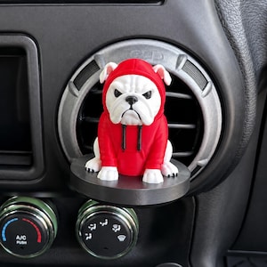 May include: A white bulldog figurine wearing a bright red hooded sweatshirt with black drawstrings. The figurine is positioned on a circular black base, which is mounted onto the horizontal slats of a vehicle air conditioning vent.