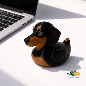 May include: A black and tan dachshund-shaped rubber duck. The duck has a black body, tan face, and ears, with a small orange accent on its tail. The duck is on a white surface near a laptop.