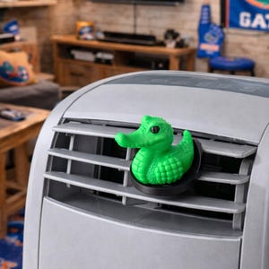 May include: A bright green alligator-shaped air freshener clip attached to the horizontal slats of a silver portable air conditioner unit. The alligator figure features a textured, scaly surface, a prominent snout, and a black circular base mount.