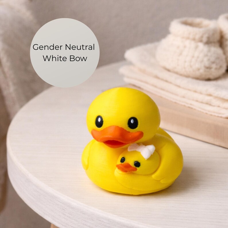 Puede incluir: Un pato de goma amarillo con pico naranja y ojos negros, que sostiene un patito m&aacute;s peque&ntilde;o con un lazo blanco. La imagen incluye el texto "Gender Neutral White Bow".