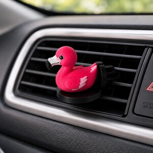May include: A bright pink flamingo car air freshener clip featuring a white and black beak, small black eyes, and white wing accents. The flamingo is attached to a circular black base designed to mount securely onto a horizontal vehicle air vent.