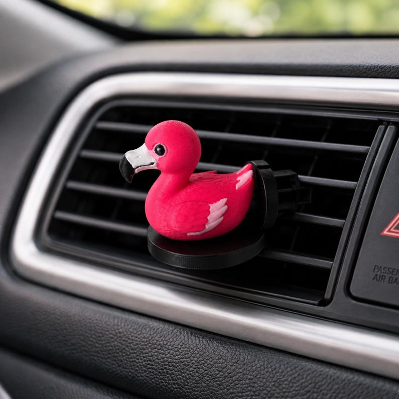 May include: A bright pink flamingo car air freshener clip featuring a white and black beak, small black eyes, and white wing accents. The flamingo is attached to a circular black base designed to mount securely onto a horizontal vehicle air vent.