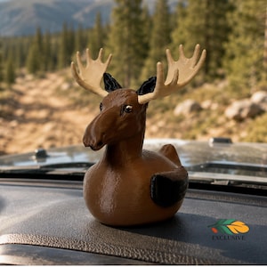 May include: A brown rubber duck-shaped figurine featuring large, light-coloured moose antlers, black ears, and black wings. The figure has a smooth, glossy finish and is positioned on a textured surface. A small logo with the word EXCLUSIVE is visible.