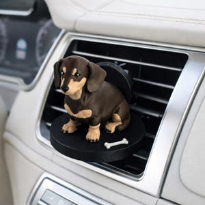 May include: A brown and tan dachshund car air vent clip-on decoration. The dog figurine sits on a black circular base with a small white bone. The air vent clip is black and the car interior is white and silver.