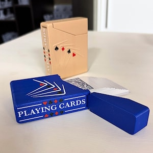 May include: A tan playing card case with a design of playing card suits and a blue playing card case with the words "PLAYING CARDS" in white. A deck of cards is visible.