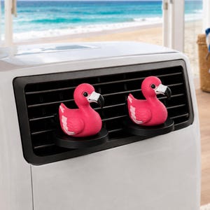 May include: Two bright pink flamingo-shaped air freshener clips attached to the black horizontal slats of a white air conditioning unit. Each flamingo features a white wing detail, a black beak, and a round black eye, mounted on a circular base.
