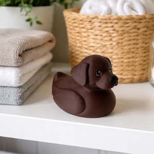 May include: A brown rubber duck shaped like a Labrador Retriever dog. The duck has a black nose, brown eyes, and floppy ears. It is sitting on a white shelf next to folded towels and a woven basket.