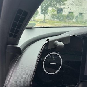 Puede incluir: Un soporte para teléfono de plástico negro mate con una rótula esférica, diseñado para engancharse al borde de la pantalla del tablero de un vehículo. El soporte está colocado cerca de un icono de interfaz digital circular con una línea.