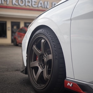 以下が含まれることがあります： 白い車の前輪とタイヤのクローズアップ。ホイールはダークブロンズ色で、マルチスポークのデザインです。タイヤは黒色で、「CONTINENTAL」の文字が見えます。車には赤いアクセントがあります。