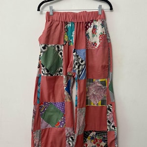Könnte beinhalten: Rosa Patchwork-Hose, die an einem schwarzen Kleiderbügel hängt. Die Hose hat eine rosa Basis mit bunten quadratischen Patches aus verschiedenen Mustern und Stoffen. Der Bund ist elastisch. Die Hose ist weit geschnitten und hat eine lockere Passform.