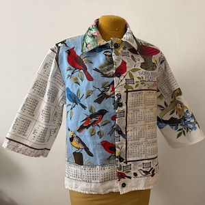 Pode incluir: Uma camisa com estampa de pássaros e calendário. A camisa tem um fundo azul claro com vários pássaros e meses do calendário. As mangas são de três quartos. O texto "SONG BIRD FEAST" é visível.