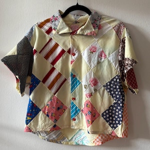 Può includere: Una camicia patchwork a maniche corte con chiusura a bottoni. La camicia presenta una varietà di quadrati di tessuto colorati, tra cui motivi floreali, a pois e a righe. Anche il colletto e i polsini sono in tessuto fantasia.